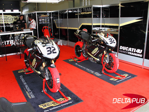 Tapis de sol publicitaires imprimés sous des motos de compétition dans un stand.